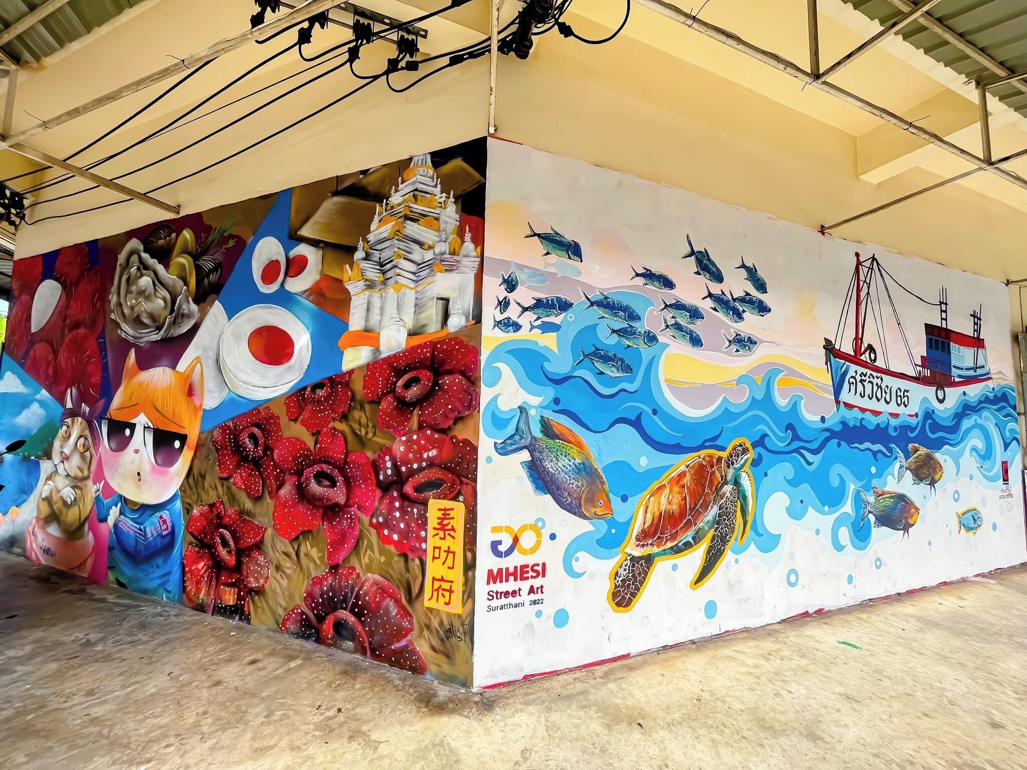 Een muurschildering met een schildpad, vissen en een bootje met de tekst 'MHESI Street Art Suratthani 2022'.