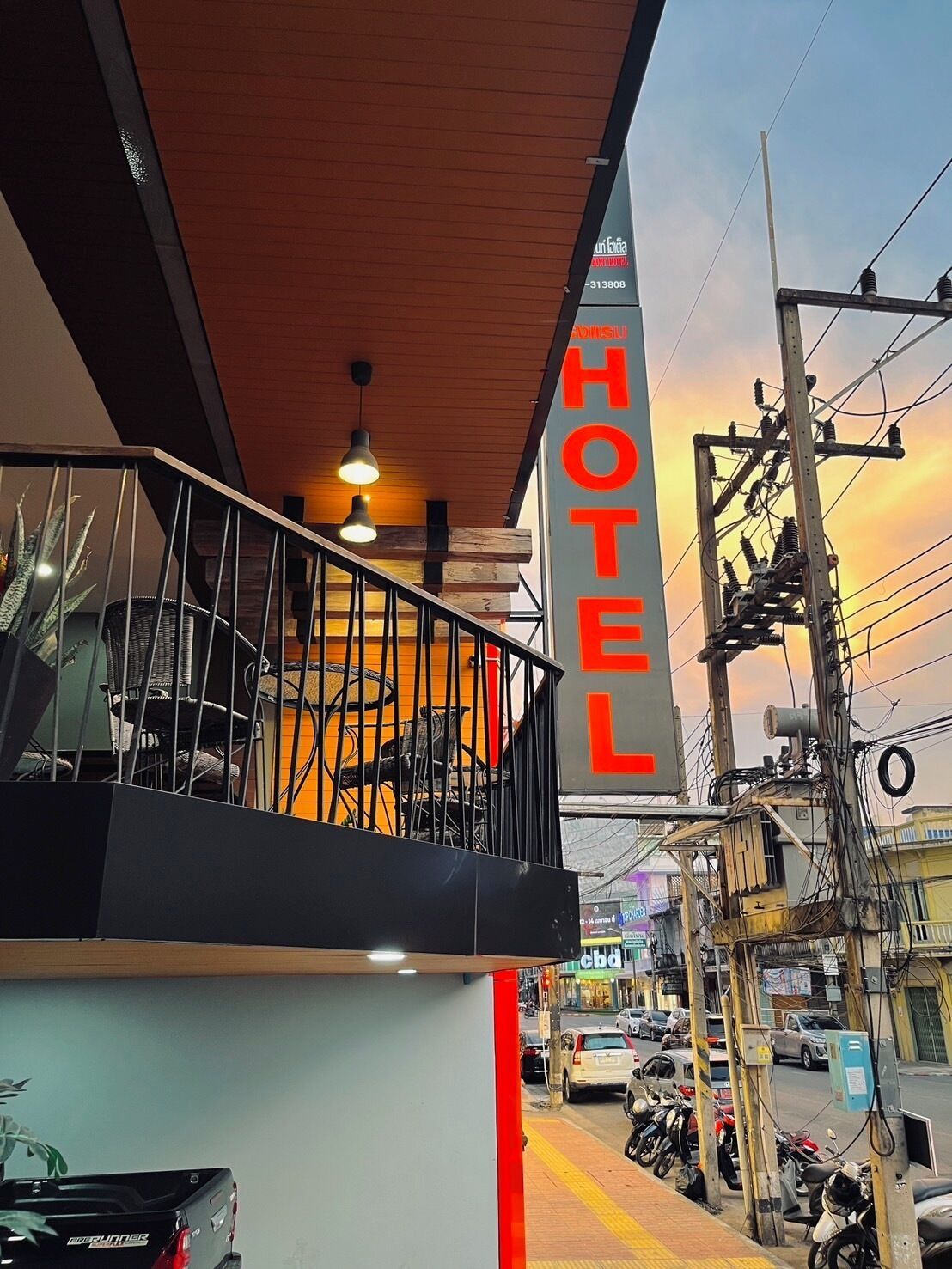 A hotel with a balcony overlooking a street, featuring a signboard with the word "HOTEL" and a view of parked cars and motorcycles below.