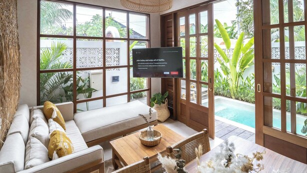 Een ruime woonkamer met een houten plafond, een groot raam met uitzicht op groen, een bank, een bijzettafeltje en een televisie.