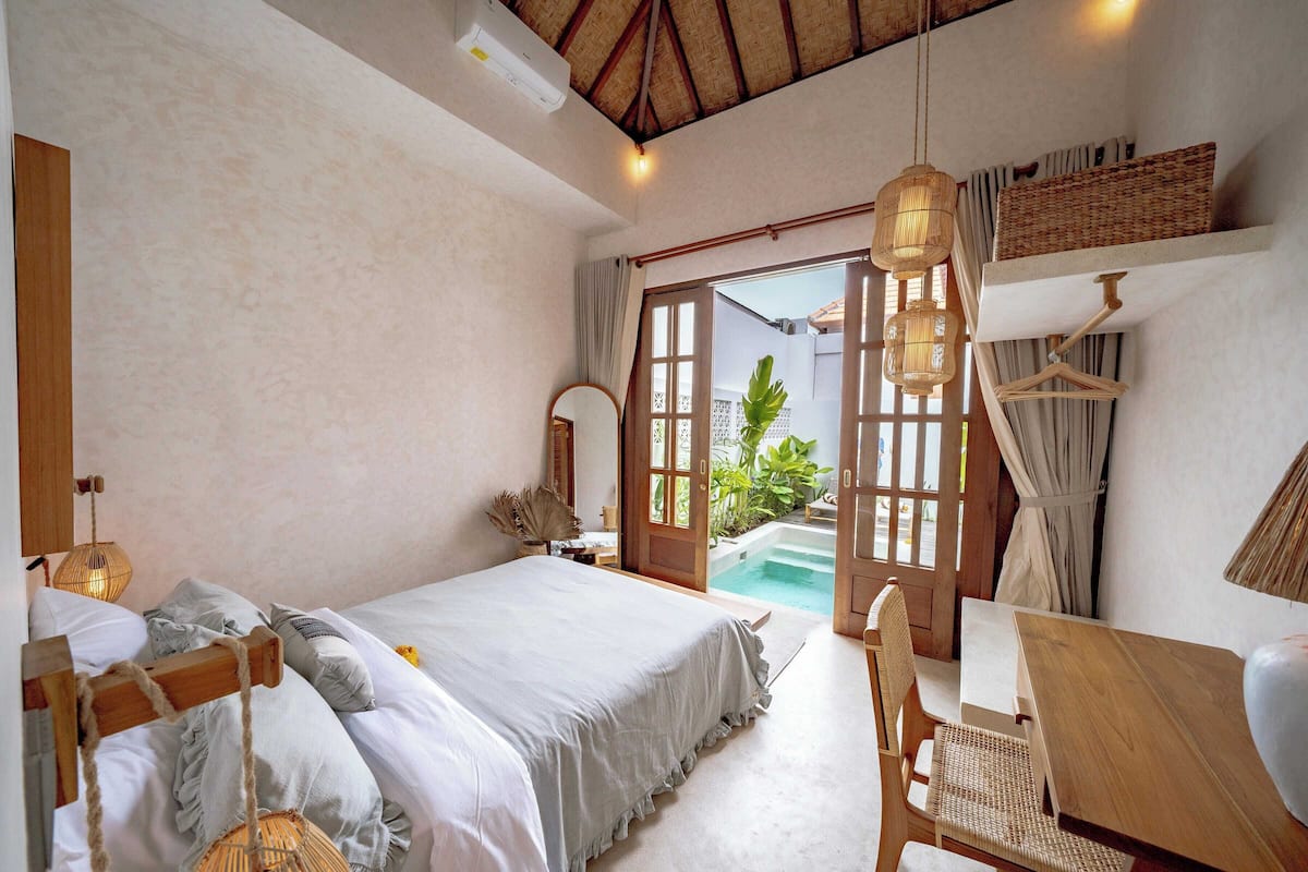 A bedroom with a bed, a wooden chair, a table, a hanging light, and a view of a pool.