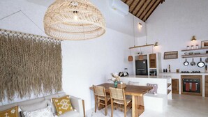 Een ruime woonkamer met houten plafond, een geweven lamp, een bank met gekleurde kussens, een houten salontafel en een keuken met moderne apparatuur.