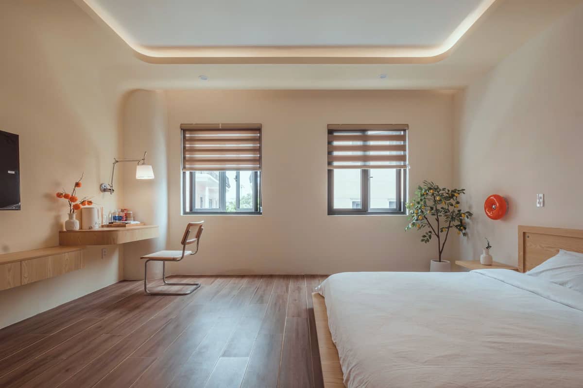 Quarto moderno com uma cama grande, uma escrivaninha com cadeira, duas janelas com persianas, um vaso de planta e uma TV fixada na parede.