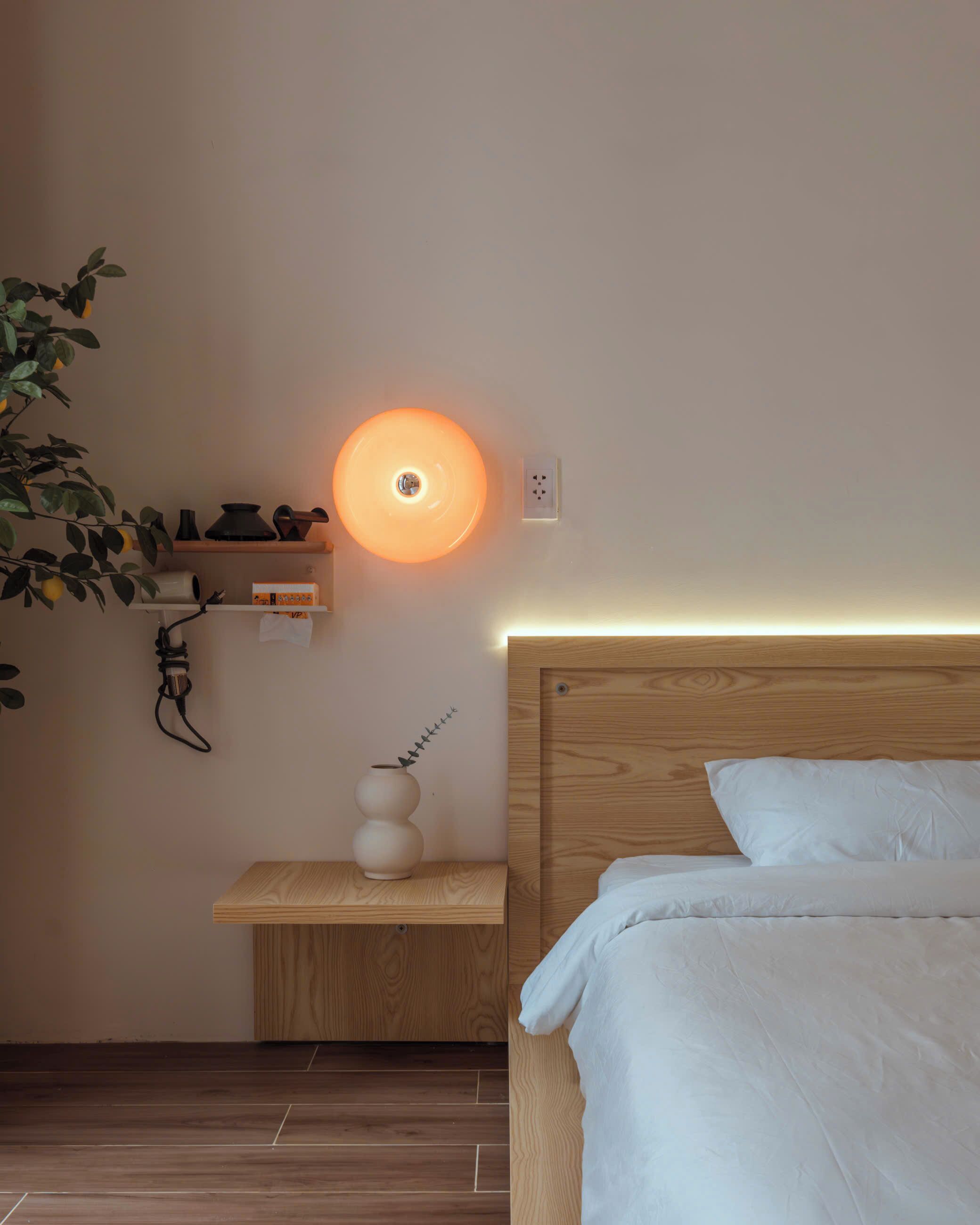 A bedroom with a wooden headboard, a bedside table with a vase, a wall-mounted shelf with decorative items, and a circular wall light.
