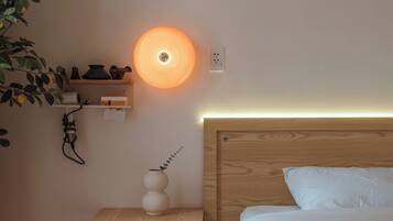 A bedroom with a wooden headboard, a bedside table with a vase, a wall-mounted shelf with decorative items, and a circular wall light.