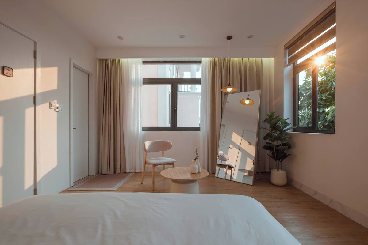 Quarto moderno com uma cama grande, uma mesa redonda de madeira, uma cadeira e um vaso com planta.