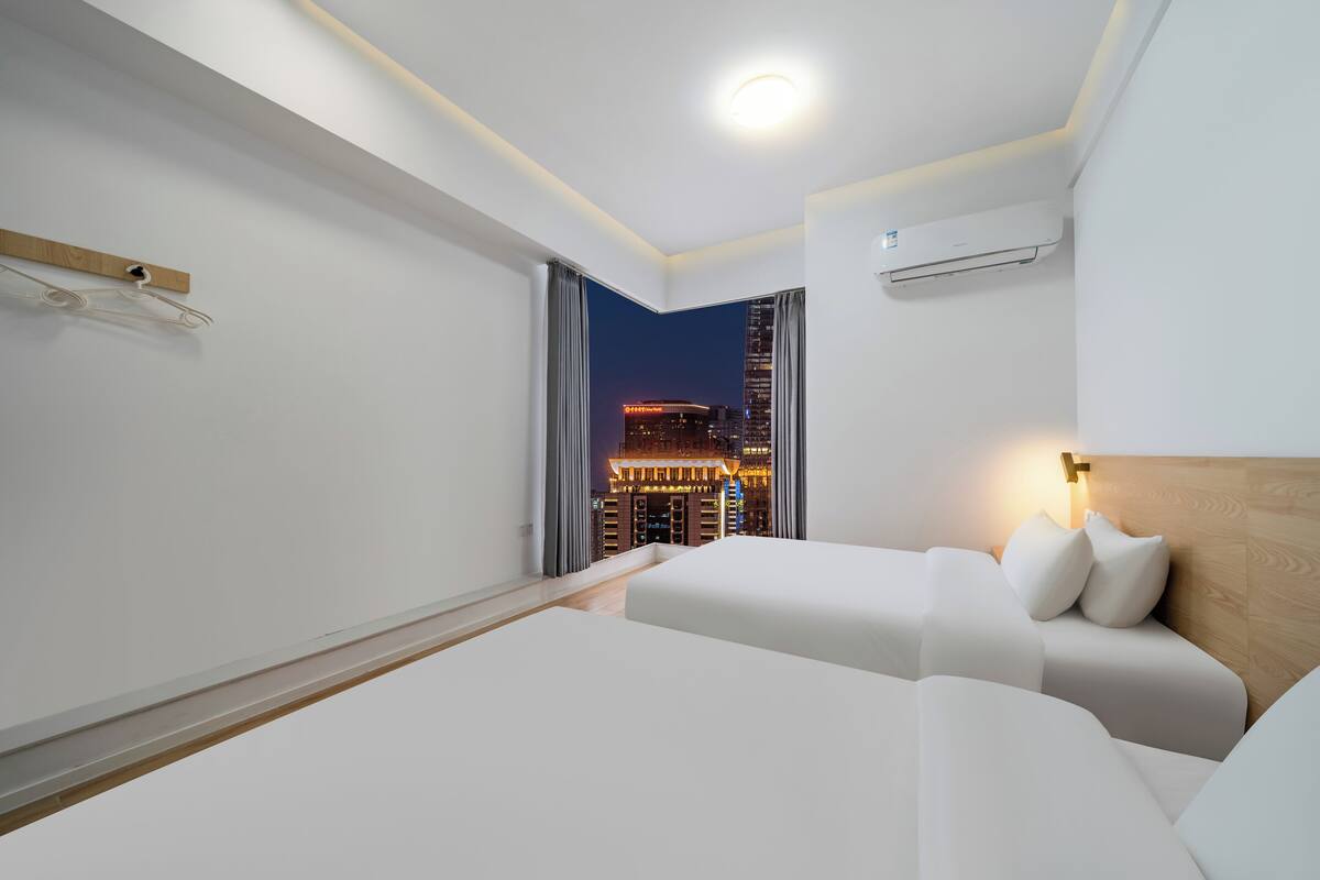 Hotelkamer met twee bedden, een houten hoofdeinde, een nachtkastje, een wandairconditioner en uitzicht op een stadsbeeld 's nachts.