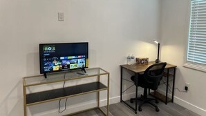 A modern room with a TV on a glass shelf, a wooden desk with a chair, and a window with blinds.