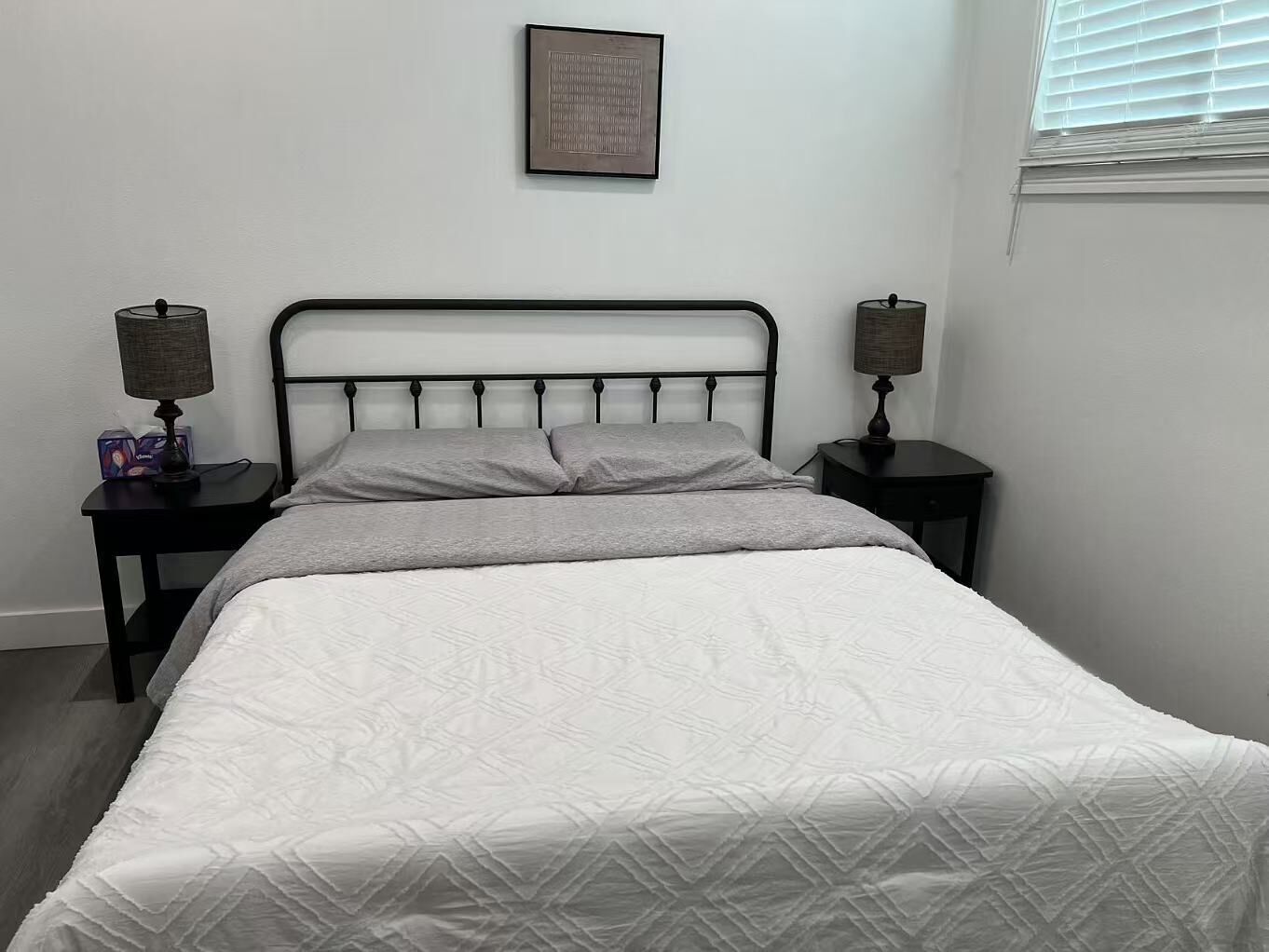 Una cama bien hecha con colcha blanca, dos mesitas de noche con lámparas y un cuadro en la pared.