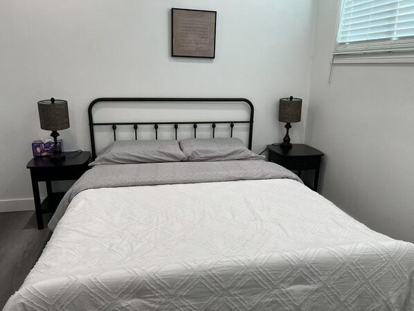 Een net opgemaakt bed met een witte deken, twee nachtkastjes met lampen en een ingelijst kunstwerk aan de muur.