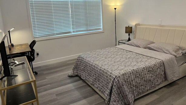 A bedroom with a bed, a desk with a chair, a standing lamp, and a window with blinds.