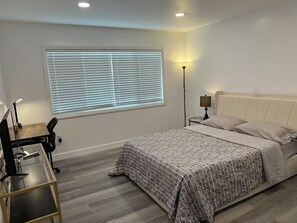 Een slaapkamer met een bed, een bureau met stoel, een staande lamp en een raam met jaloezieën.
