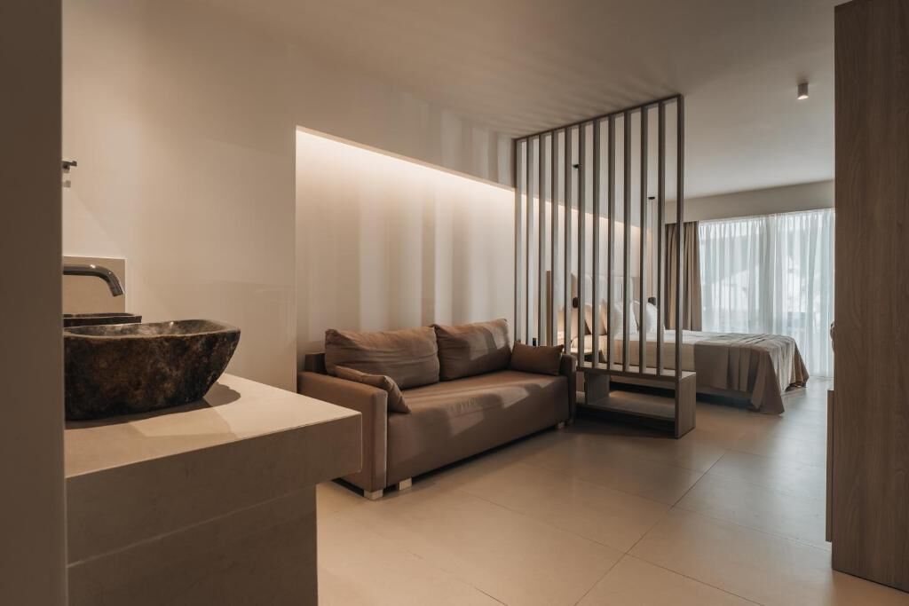 A modern hotel room with a large stone sink, a beige sofa, a wooden crib, and a large window with sheer curtains.