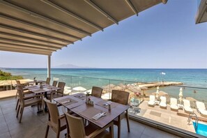 A terrace with tables and chairs overlooking a beach and the sea. - Golden Coast Suites (Zakynthos)