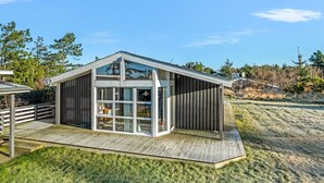 Une cabane moderne en bois, dotée d’une grande porte et de grandes fenêtres en verre, d’une terrasse et d’un toit à pente.