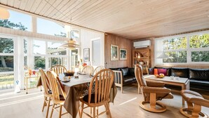 Une pièce spacieuse avec un plafond en bois, de grandes fenêtres, un coin repas avec des chaises et une table en bois, un canapé en cuir noir et une bibliothèque.