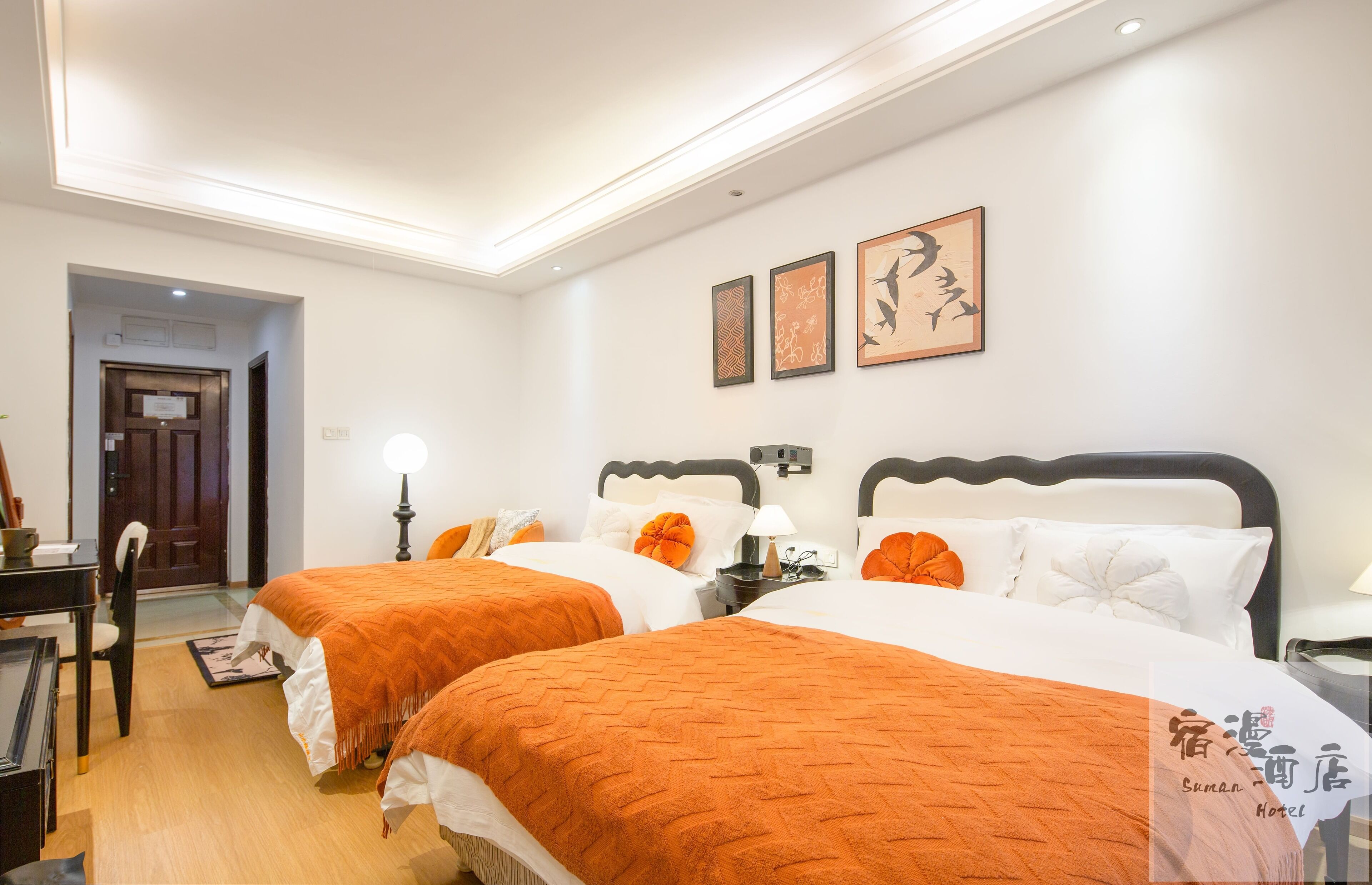 A hotel room with two beds, each with an orange bedspread, white pillows, and a black headboard. There is a desk with a chair, a lamp, and a framed picture on the wall.