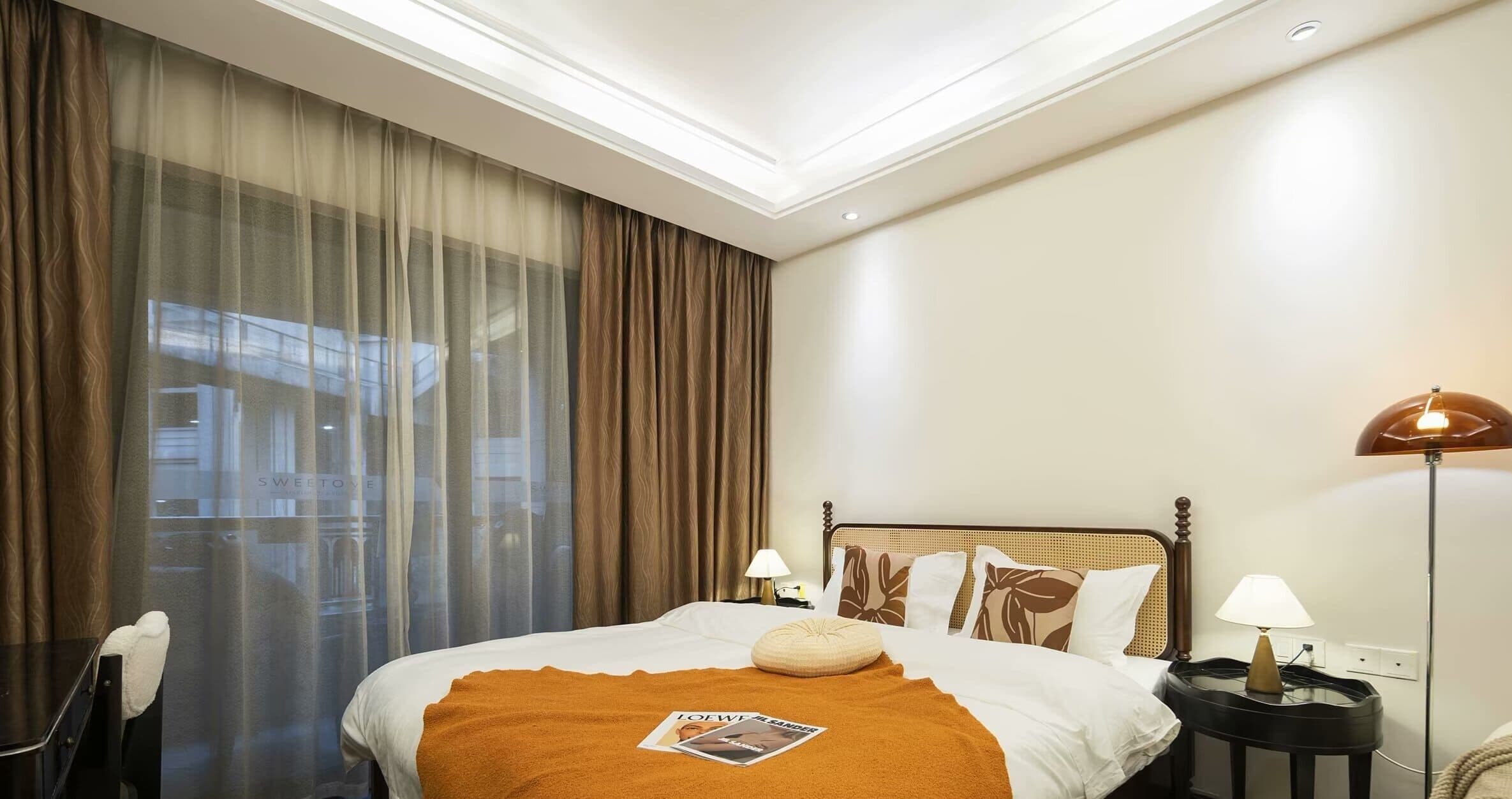 A hotel room with a large bed, a nightstand, a lamp, and a view of the outside through sheer curtains.