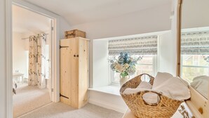 Ein Zimmer mit einem Holzschrank, einem Korbwäscheschrank mit Handtüchern, einem Fenster mit Blick auf Gebäude und einer Vase mit Blumen.