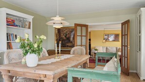 Un comedor con una mesa de madera, sillas de mimbre, un jarrón blanco con flores y una estantería.