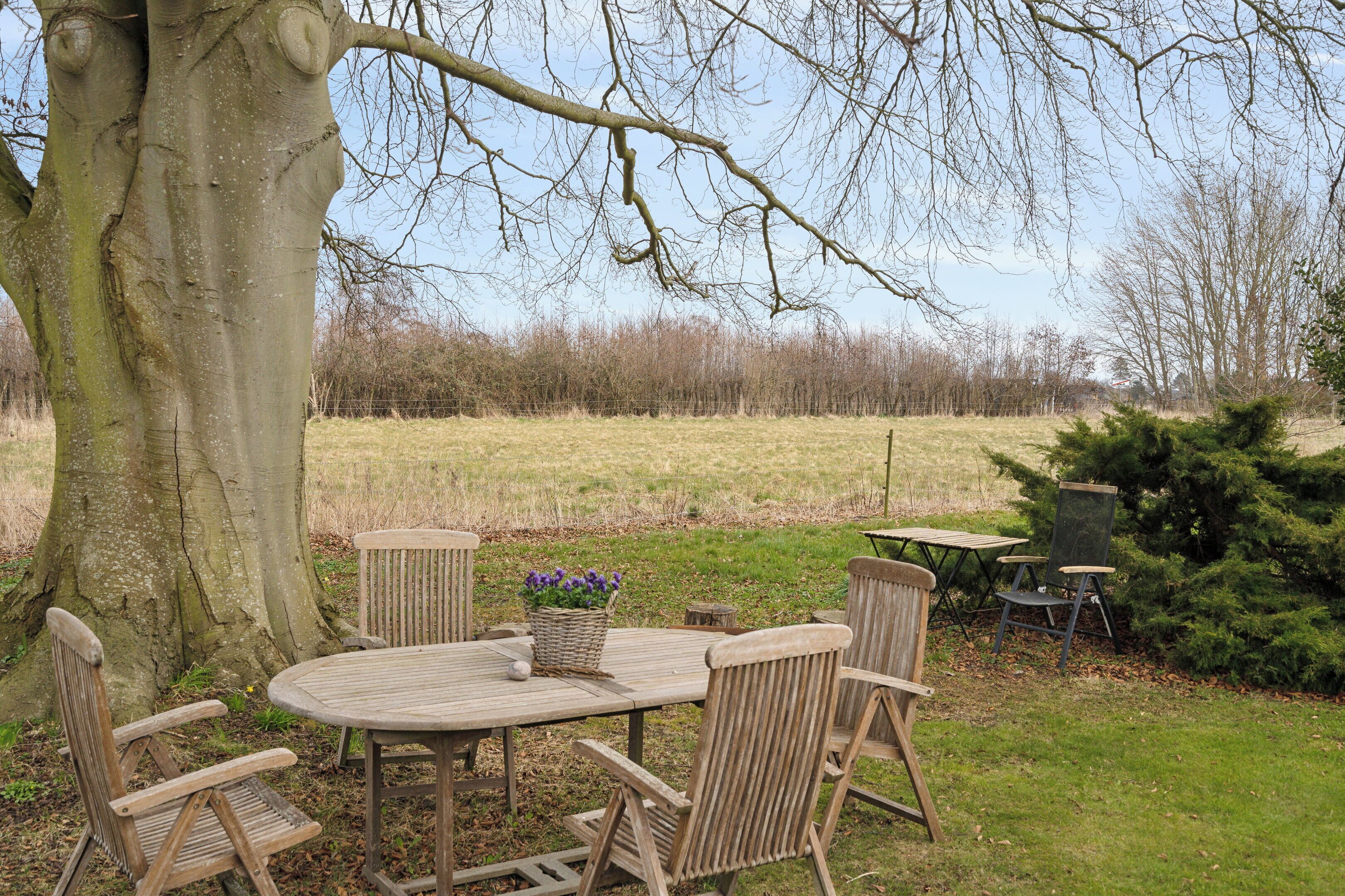 Un espace extérieur aménagé avec une table ronde en bois et des chaises, une petite table avec une chaise, et un arbre en arrière-plan.