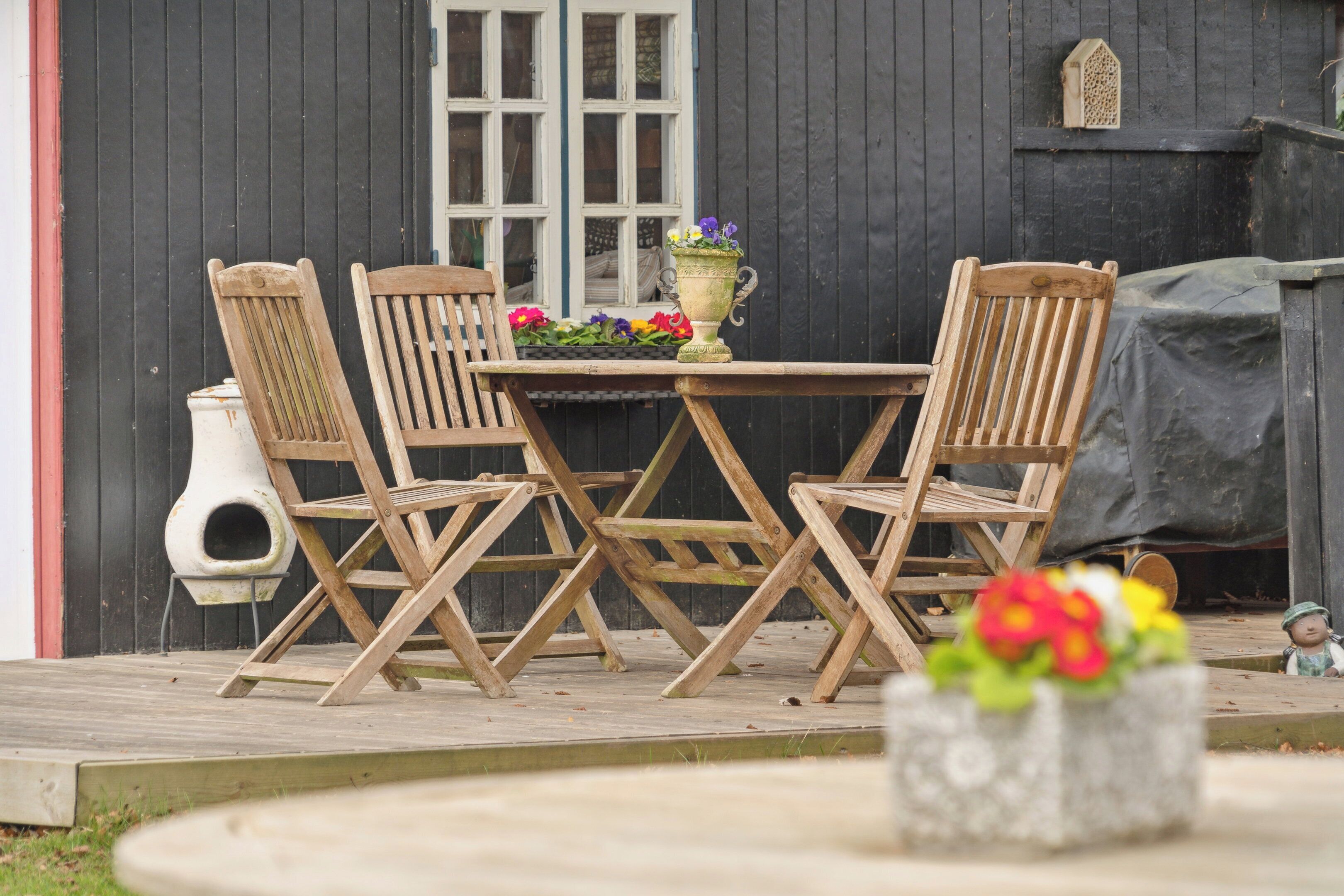 A wooden table and chairs set outdoors with a window and a birdhouse in the background.