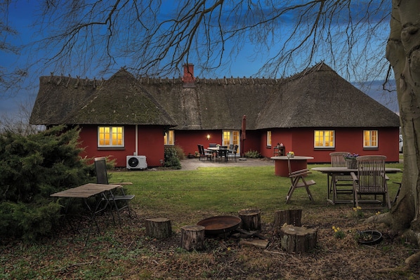 Een rieten huis met een rode gevel, een terras met tuinmeubels en een verzorgd gazon.