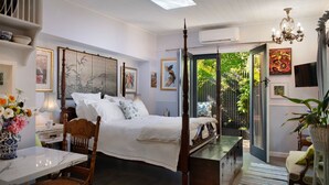 Un dormitorio con una cama con dosel, una mesita de noche con lámpara, una silla y una pequeña mesa con un florero. Hay cuadros en la pared y una ventana con vistas a la vegetación.