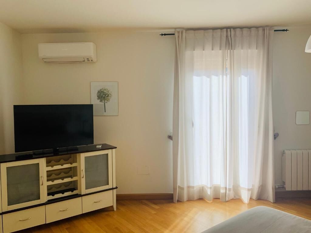 Una habitación con televisión de pantalla plana sobre un mueble, un radiador y cortinas de encaje.