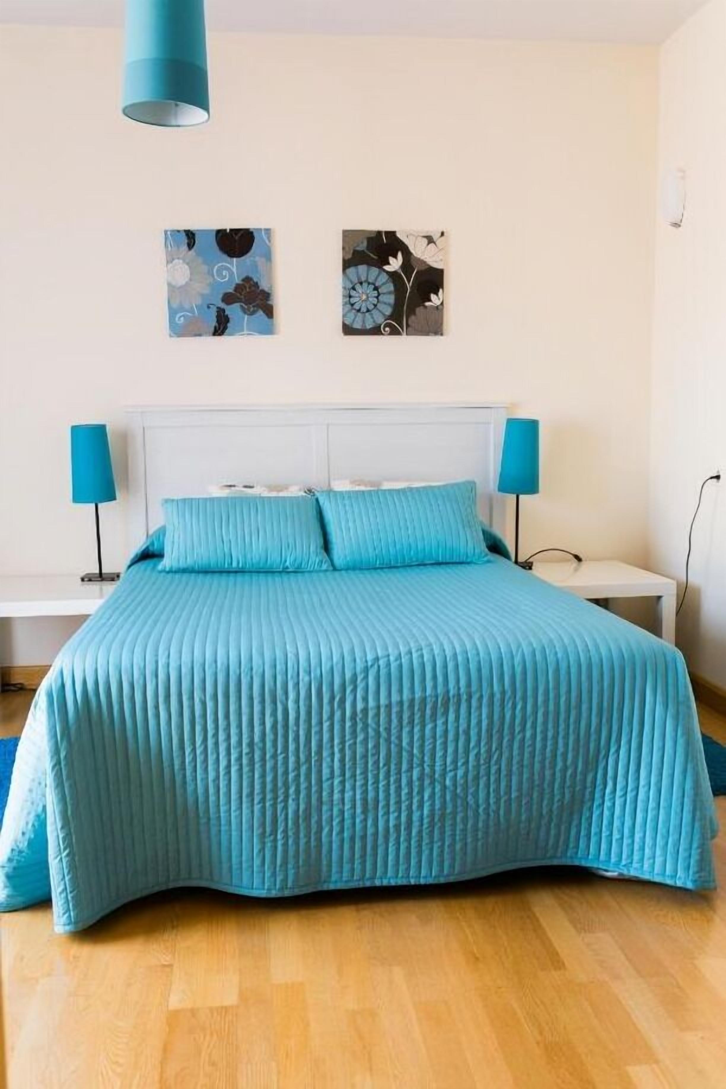 A bedroom with a bed, two wall-mounted lamps, and two framed artworks.
