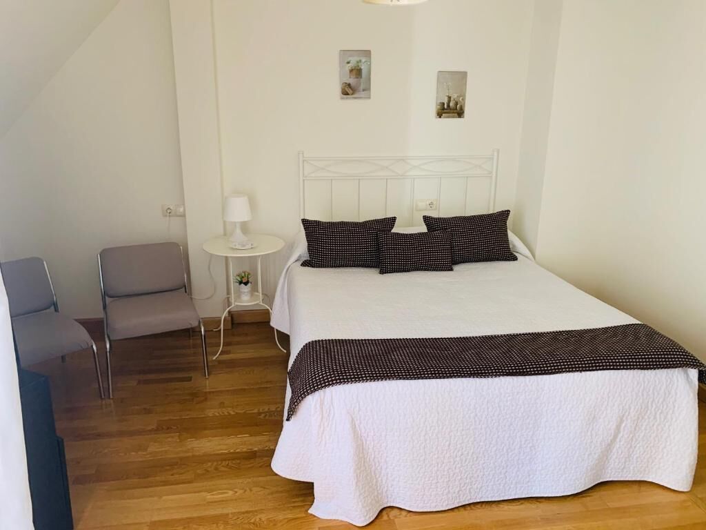 A bedroom with a white bed, two pillows, a bedside table with a lamp, and a chair.