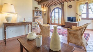 A cozy living room with a wooden table, wicker chairs, a TV, and a large window with curtains. - Casa Viola Pool, Wifi, Montaione (Montaione)