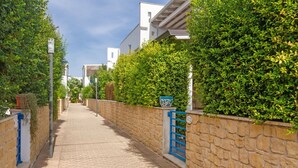 A paved pathway flanked by stone walls and lush greenery, leading to a building with a blue gate.