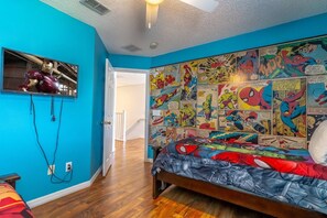 Une chambre avec un mur d’un bleu vif, un lit recouvert d’une couette aux motifs de Spider-Man, et une étagère murale pour exposer des bandes dessinées.