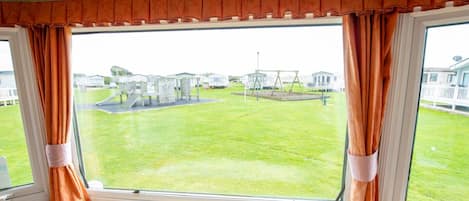 A room with a large window, curtains tied back, and a view of a grassy outdoor area with playground equipment.