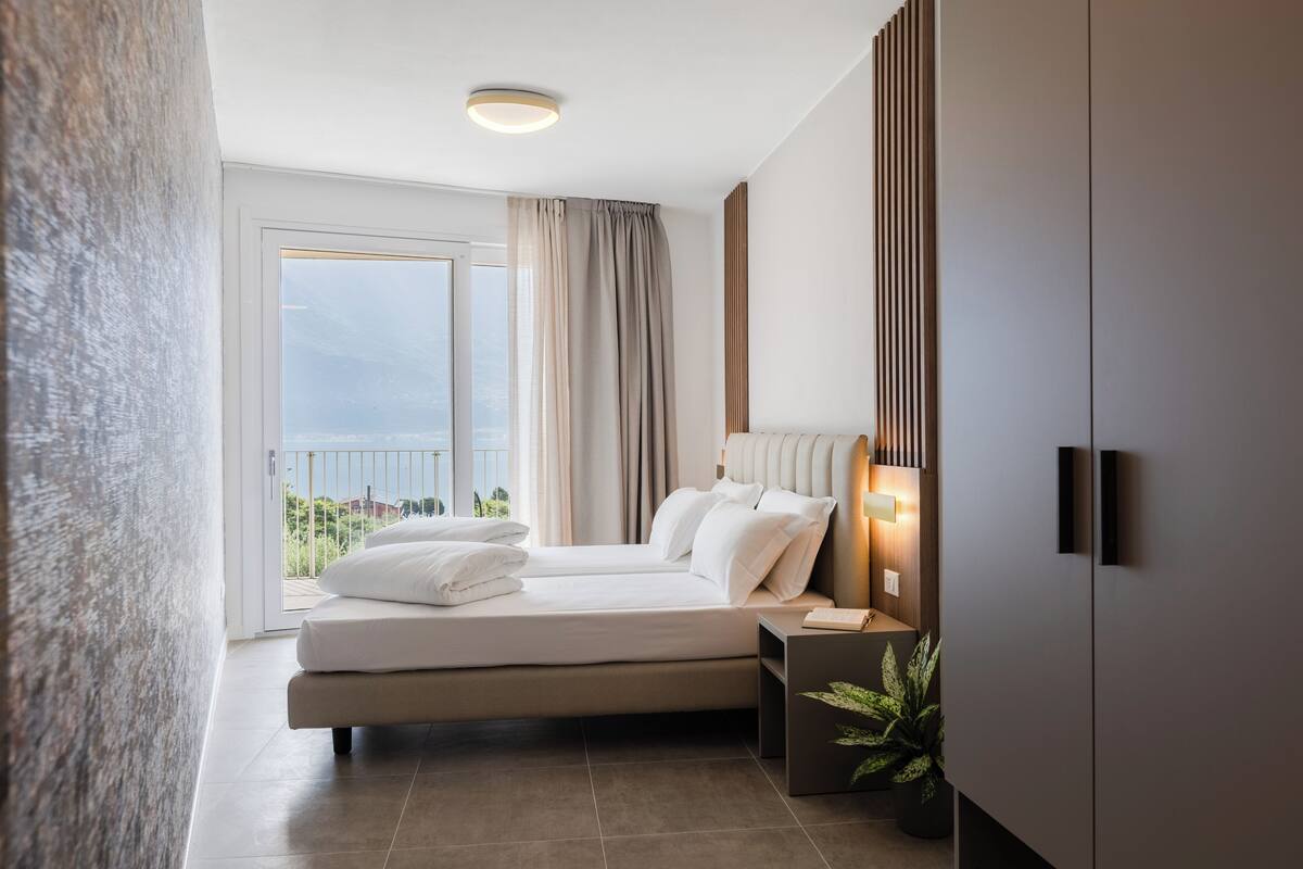 Una camera da letto moderna con un letto grande, un comodino con una lampada, una pianta in vaso e vista sull'esterno.
