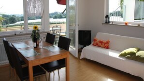 A room with a dining table, chairs, a sofa, a TV, and large windows with a view of greenery. - Apartment "Panorama" with Lake and Mountain Views, Wi-Fi (Friedrichshafen)