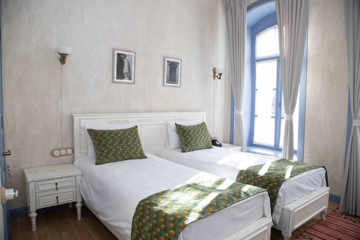 Una cama doble con cabecera blanca, dos mesitas de noche y una ventana con cortinas.