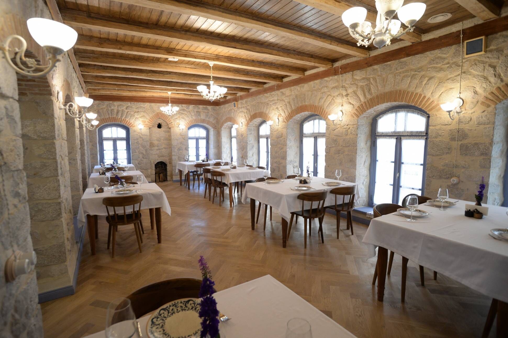 Ein Restaurant mit Steinwänden, Holzbalken und Rundbogenfenstern. Die Tische sind mit weißen Tischdecken gedeckt und es stehen Stühle bereit.