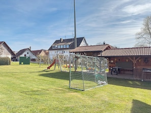 Un espacio al aire libre con césped, un parque infantil con tobogán y columpios, y una portería de fútbol.