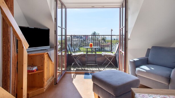 A room with a TV, a sofa, a balcony with a table and chairs, and a wooden cabinet. - Haus Hilebrand - Apartment '4, 75 sqm' with Shared Garden, Balcony and Wi-Fi (Kressbronn a. B.)