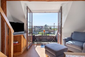 A room with a TV, a sofa, a balcony with a table and chairs, and a wooden cabinet. - Haus Hilebrand - Apartment '4, 75 sqm' with Shared Garden, Balcony and Wi-Fi (Kressbronn a. B.)