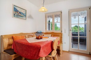 Un coin repas avec un banc en bois, une table recouverte d’une nappe rouge, un tableau accroché au mur et une fenêtre ornée de rideaux en dentelle.