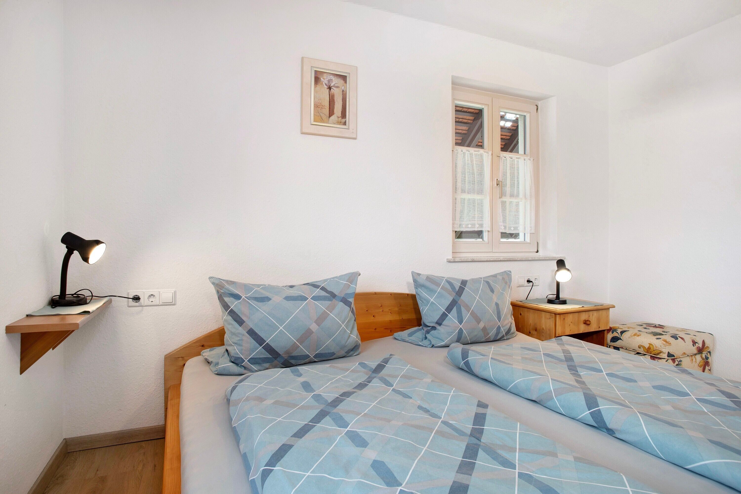 A bedroom with a bed, two bedside tables with lamps, a window, a wall-mounted shelf, and a picture.