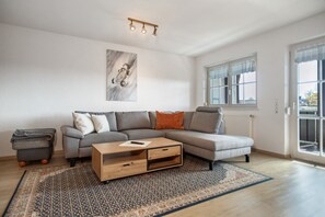 Un salon moderne avec un canapé d’angle, une table basse en bois, un tapis à motifs et une grande fenêtre avec des rideaux.