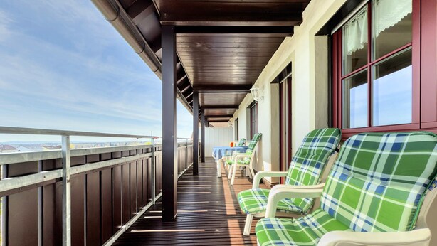 A covered outdoor seating area with wooden flooring and white plastic chairs with green and blue checkered cushions.