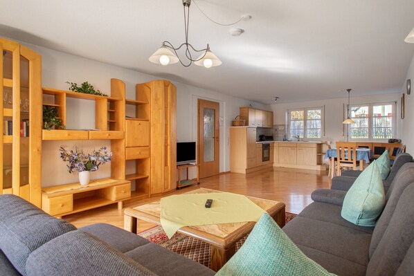 A spacious living room with a sofa, wooden shelving, a television, and a kitchen area in the background. - Haus Hilebrand – Apartment '1, 80 sqm' with Shared Garden, Balcony and Wi-Fi (Kressbronn a. B.)