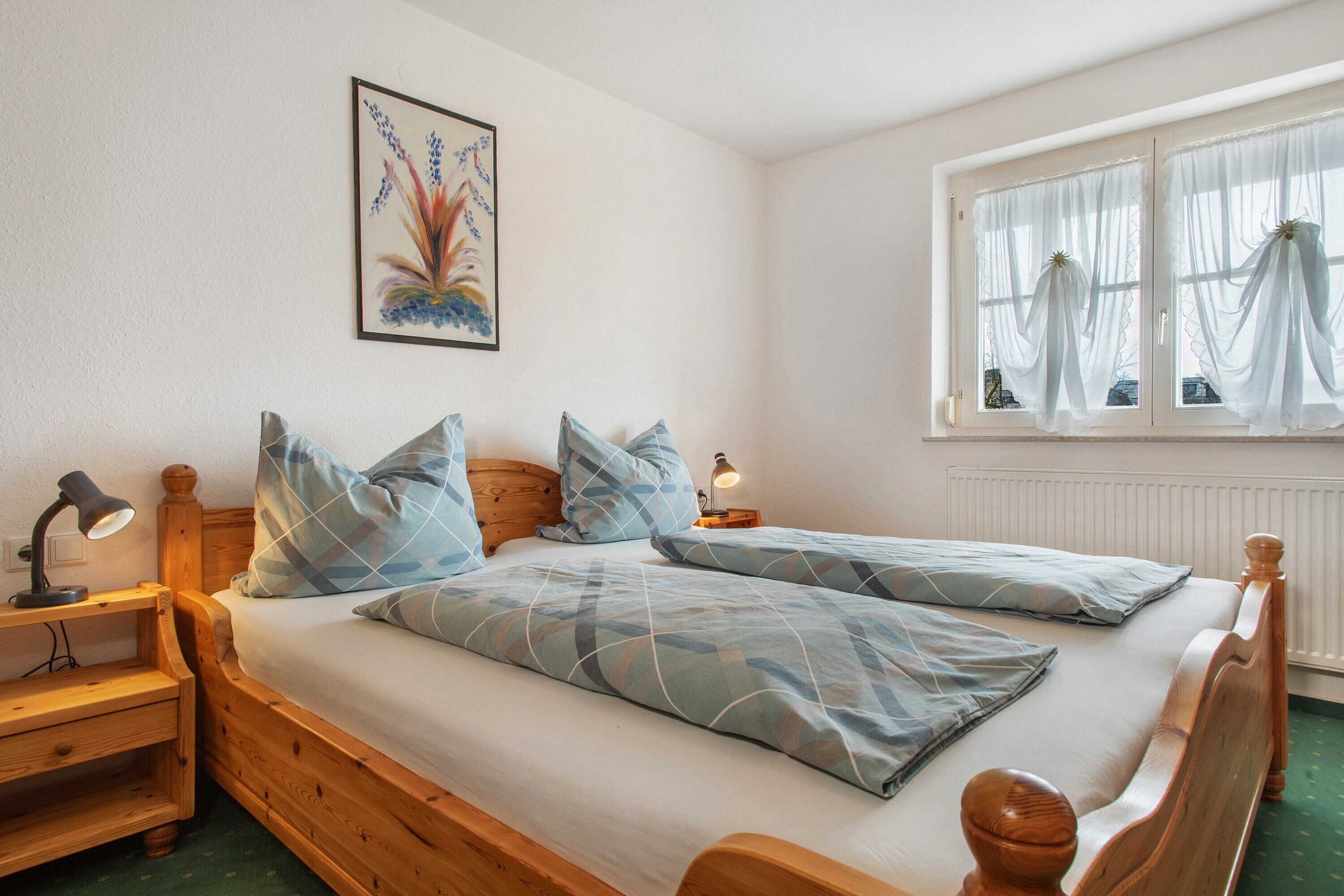 A double bed with a wooden frame, two bedside tables with lamps, a window with sheer curtains, and a wall-mounted painting.