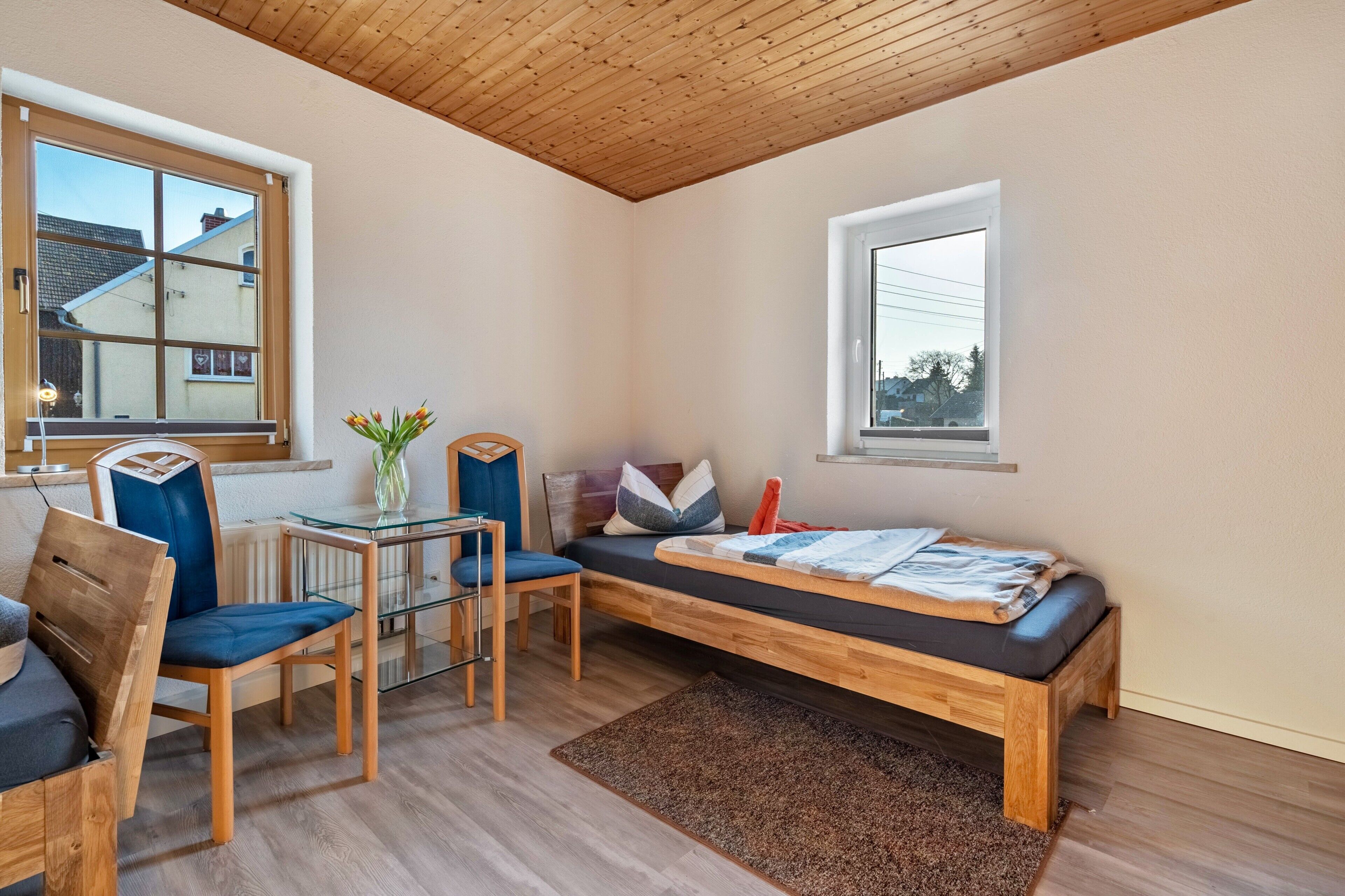 A compact room with a bed, a chair, a table, and two windows.