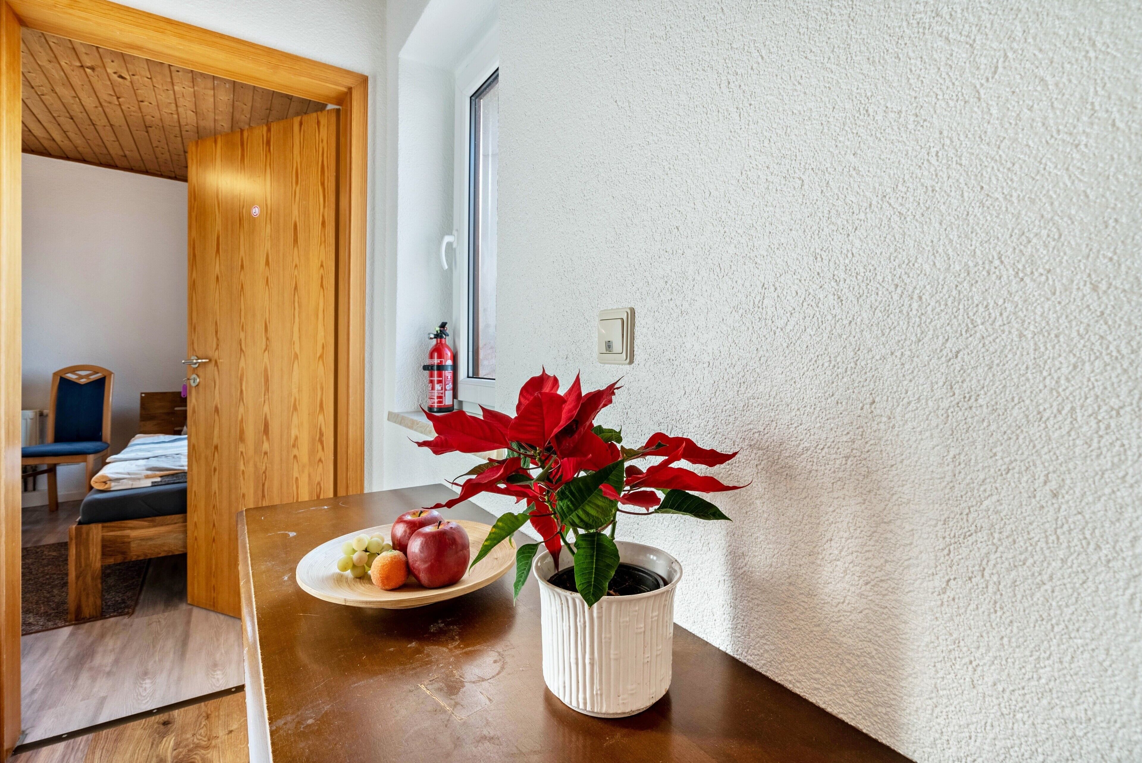 A wooden dining table with a white pot containing a red plant and a plate of fruit.