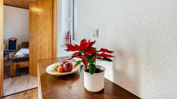 A wooden dining table with a white pot containing a red plant and a plate of fruit.